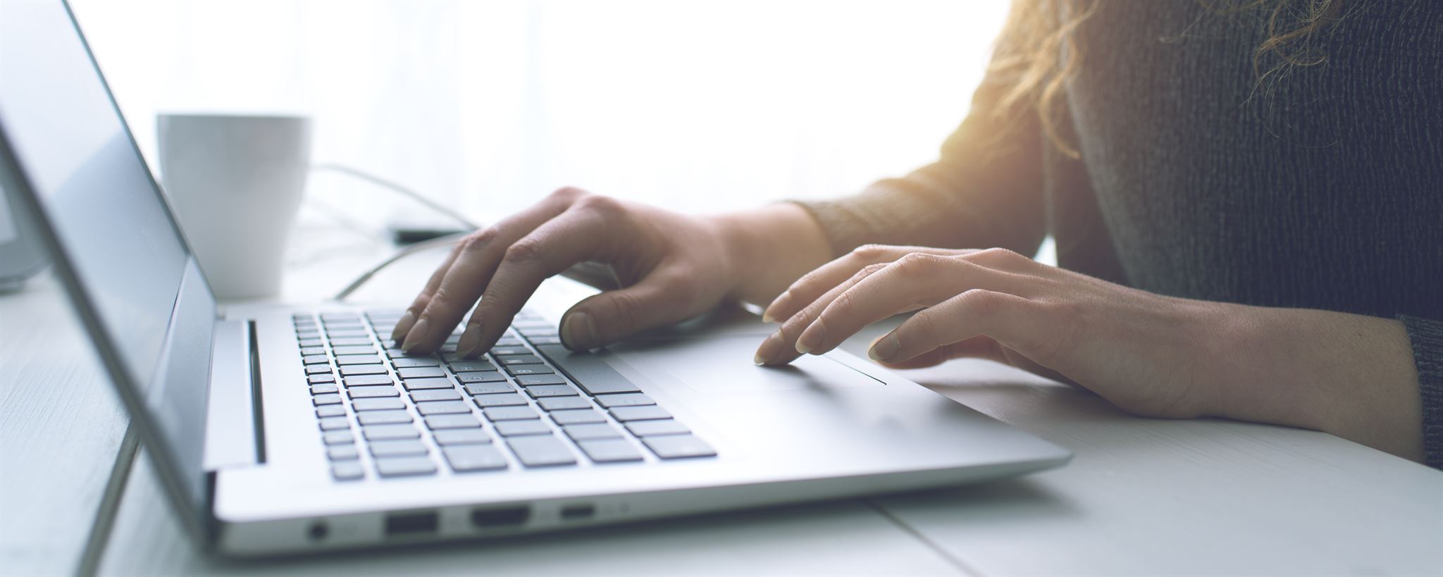 Close-up-of-womans-hands-typing-and-using-a-trackpad-on-a-laptop-with-mug